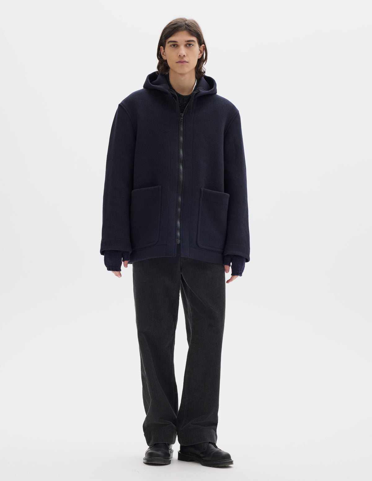 Full body image, front view of model wearing a dark navy zip through wool coat, with a hood, two laid-on patch pockets, straight cuffs and hems. Worn over a dark grey wool jumper and styled with navy fingerless gloves, dark grey cord trousers and black leather shoes.