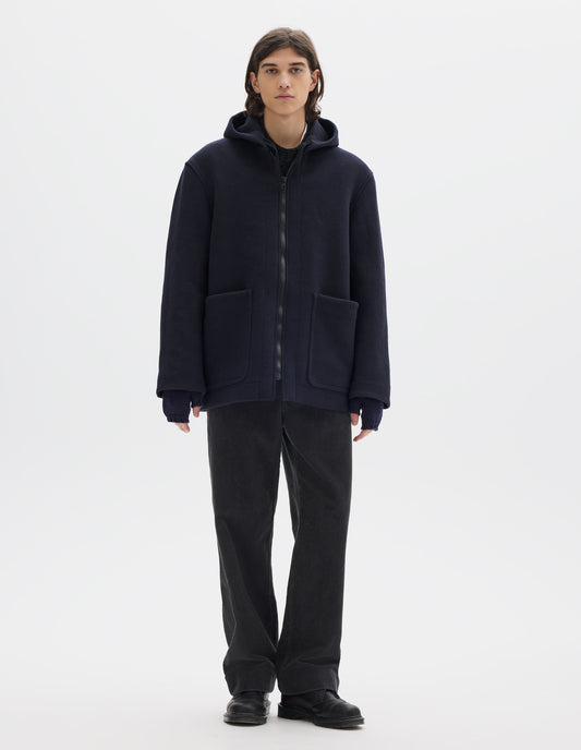 Full body image, front view of model wearing a dark navy zip through wool coat, with a hood, two laid-on patch pockets, straight cuffs and hems. Worn over a dark grey wool jumper and styled with navy fingerless gloves, dark grey cord trousers and black leather shoes.