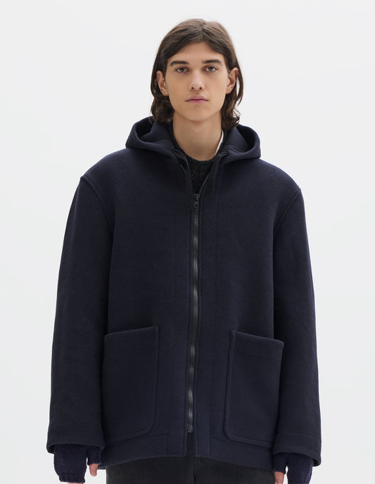 Cropped body image, front view of model wearing a dark navy zip through wool coat, with a hood, two laid-on patch pockets, straight cuffs and hems. Worn over a dark grey wool jumper and styled with navy fingerless gloves and dark grey cord trousers.