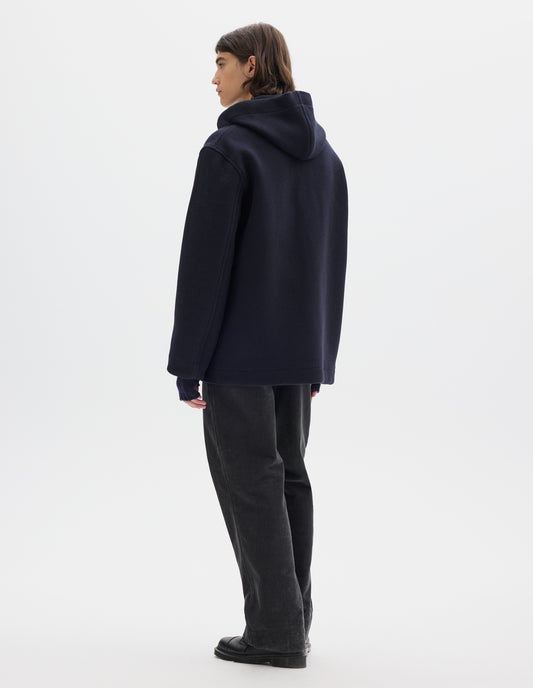 Full body image, back view of model wearing a dark navy wool coat, with a hood and straight cuffs and hems. Styled with navy fingerless gloves, dark grey cord trousers and black leather shoes.