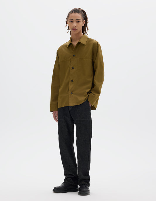 Full body image, front view of model wearing a cotton poplin green shirt with stitch through placket and pocket insert, grown-on cuffs and dark corozo buttons. Styled with black denim trousers and black leather shoes.