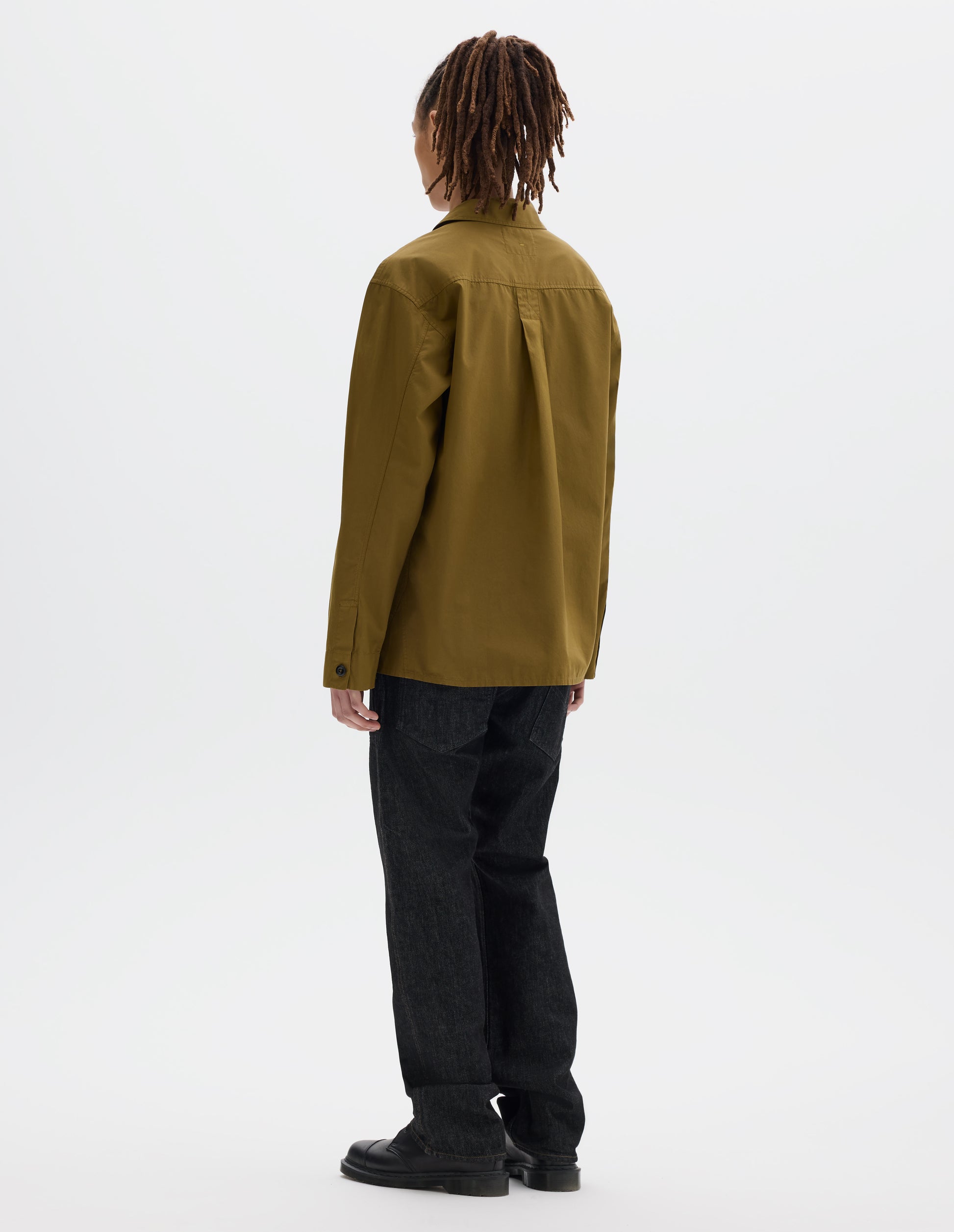 Full body image, back view of model wearing a cotton poplin green shirt, a box pleat on the back yoke and grown-on shirt cuffs, finished with dark corozo button. Styled with black denim trousers and black leather shoes.