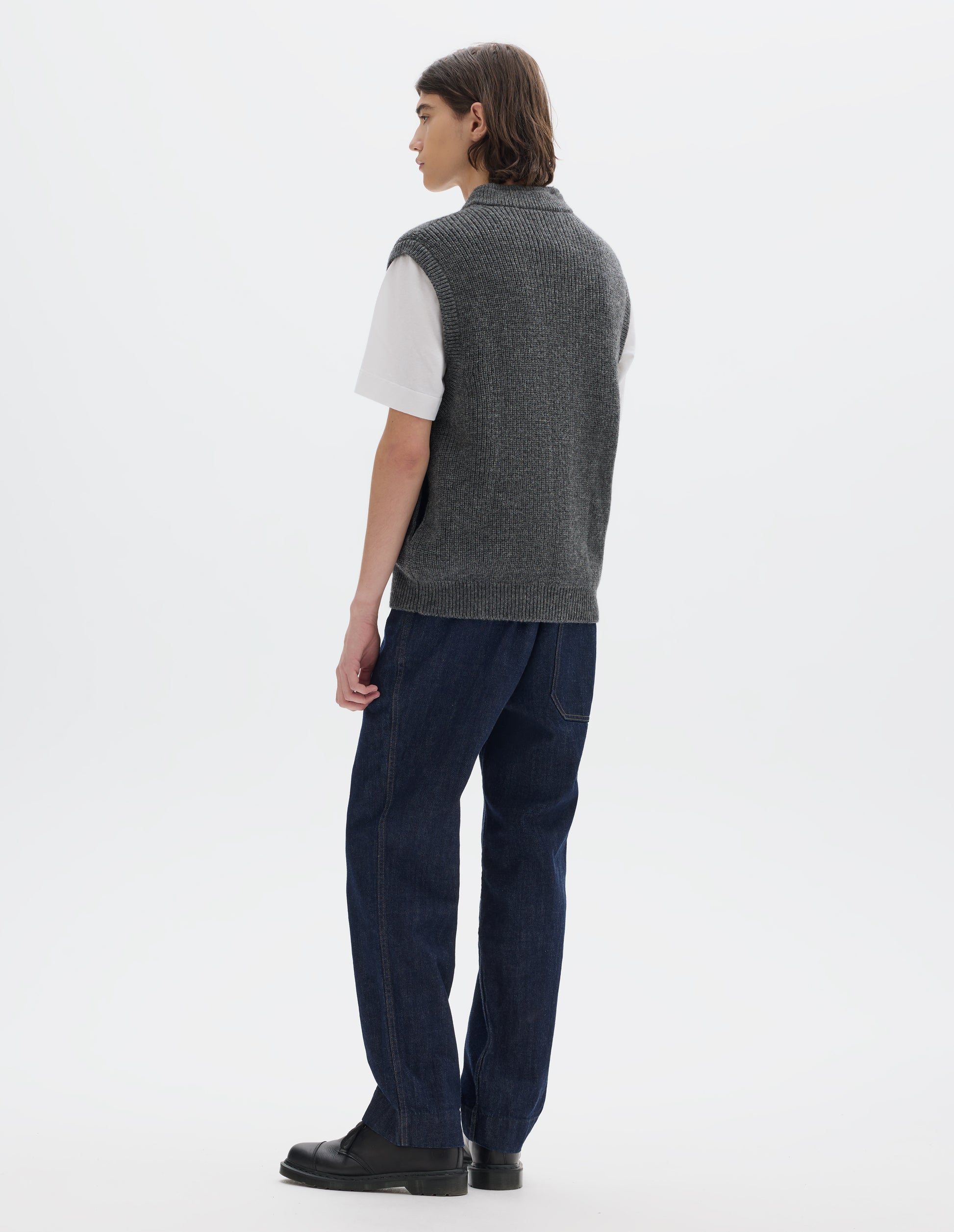 Full body image, back view of model wearing grey knitted wool sleeveless gilet, with rib neck, armholes and hem. Styled over white t shirt with blue denim jeans and black leather shoes.