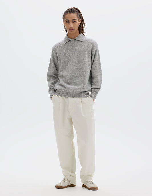Full body image, front view of model wearing a light grey wool collared sweater  with a wide rib linked on collar, plain knit body, and deeper ribbed cuffs. Styled with white pleated trousers and off white suede trainers.
