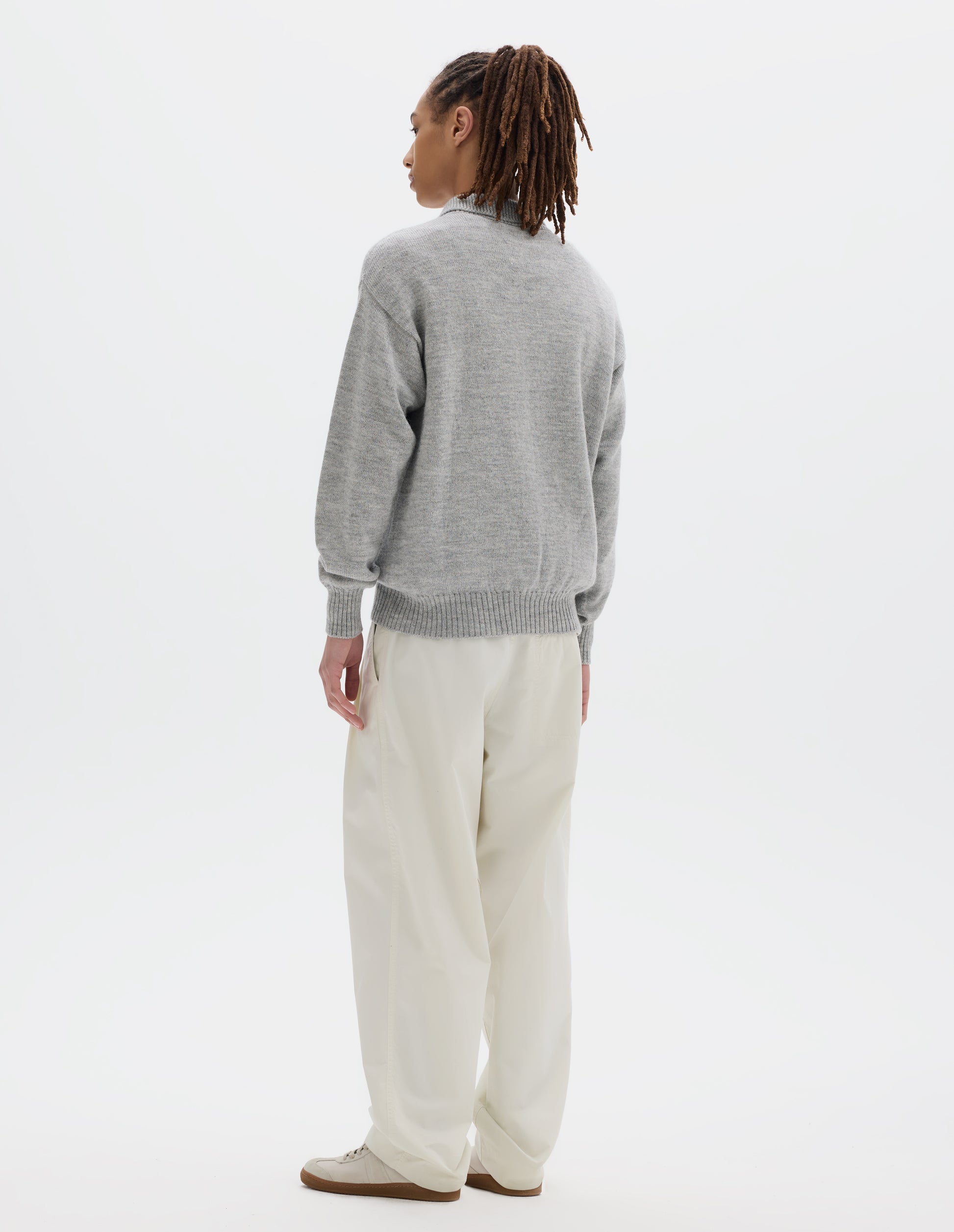 Full body image, back view of model wearing a black wool collared sweater  with a wide rib linked on collar, plain knit body, and deeper ribbed cuffs. Styled with white trousers and off white suede trainers.