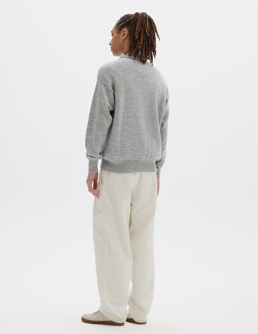 Full body image, back view of model wearing a black wool collared sweater  with a wide rib linked on collar, plain knit body, and deeper ribbed cuffs. Styled with white trousers and off white suede trainers.