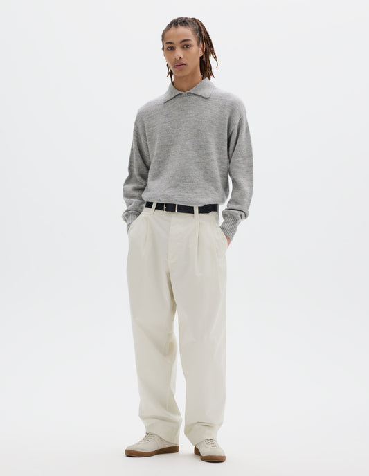 Full body image, front view of model wearing a light grey collared knitted jumper, styled with a black leather belt and an off white double pleat cotton trousers with a stitch-through, button-fastening waistband. Model is wearing pale beige trainers.