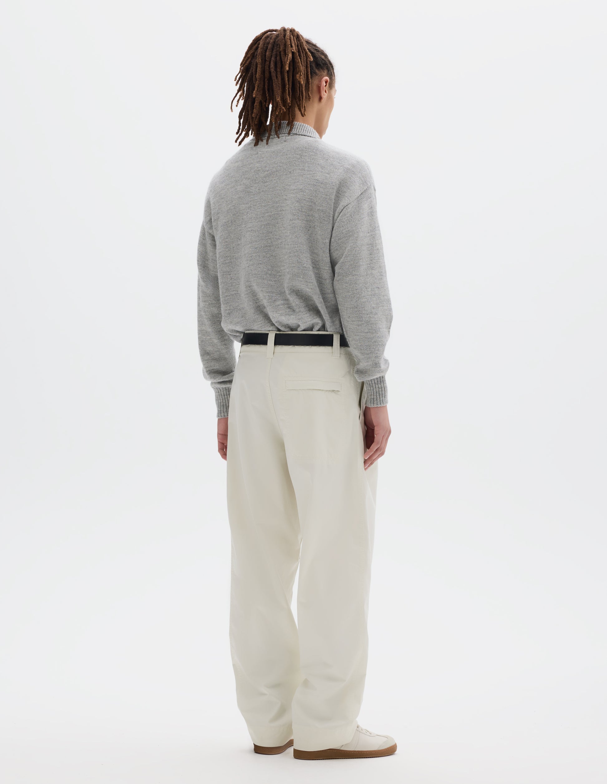 Full body image, back view of model wearing a light grey collared knitted jumper, styled with a black leather belt and an off white pleated cotton trousers with a stitch-through back pocket with a concealed zip fastening. Model is wearing pale beige trainers.