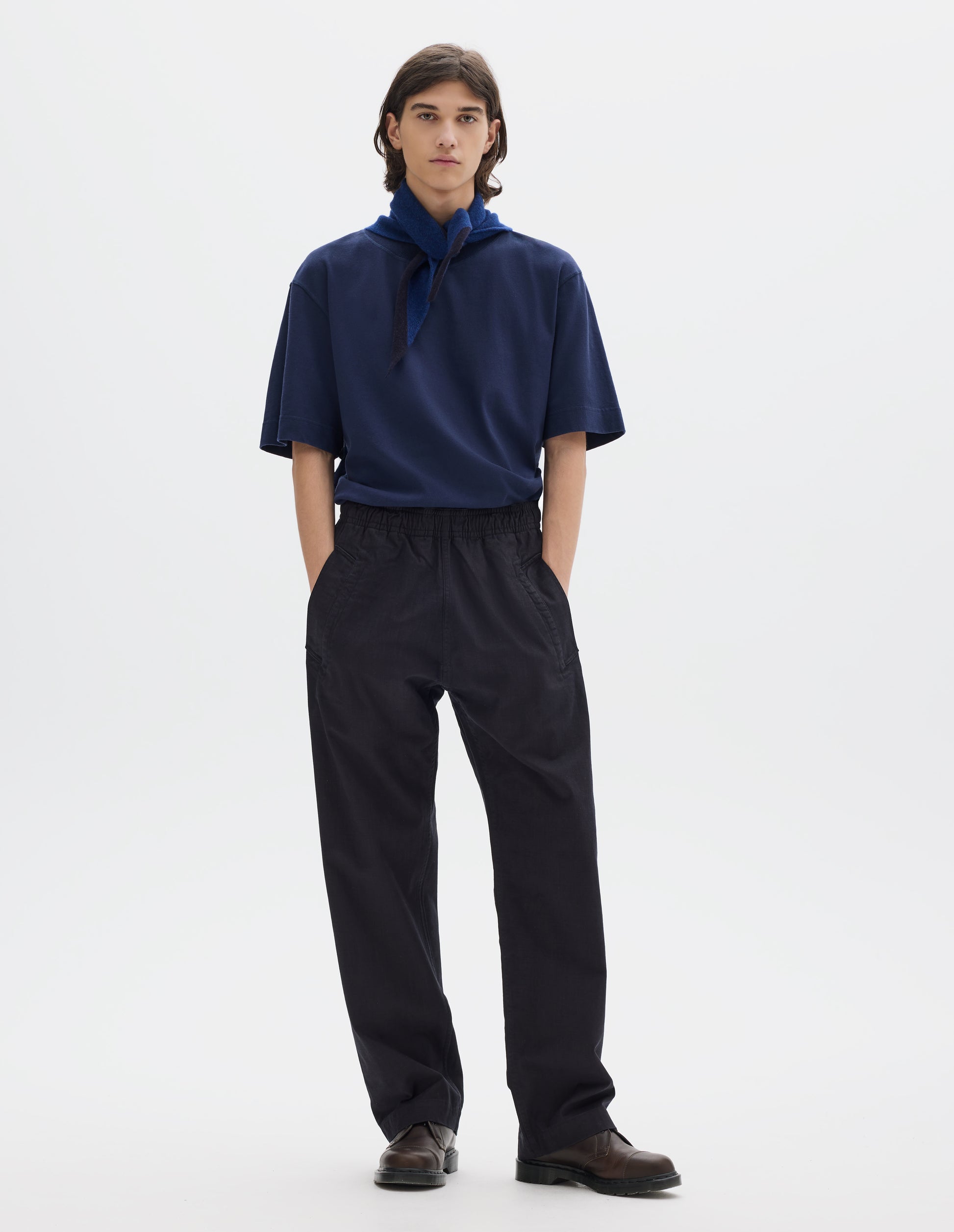 Full body image, front view of model wearing relaxed dark blue joggers with elasticated waist and deep welt pockets. Styled with a relaxed blue short sleeve t-shirt, blue and navy wool scout scarf and dark brown leather shoes.