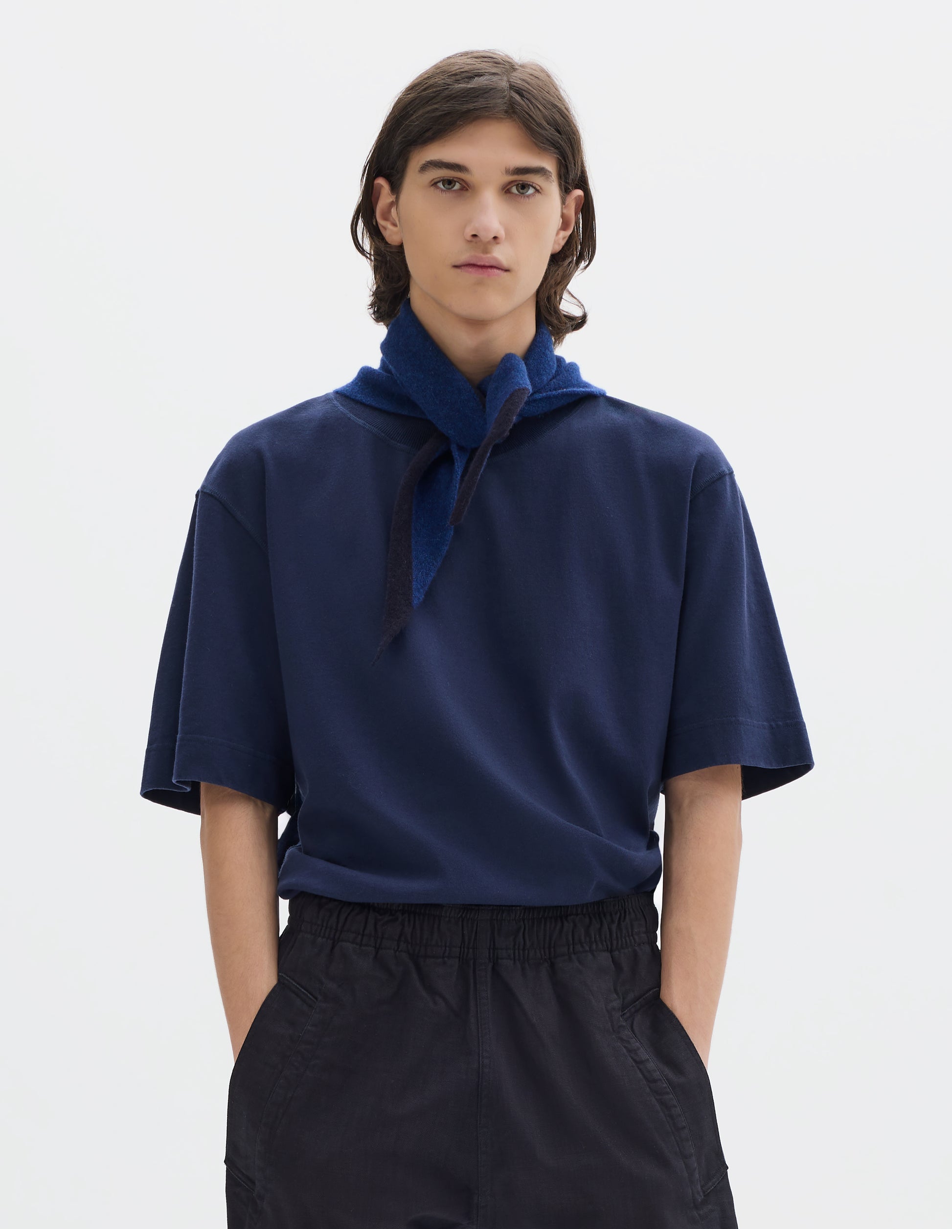 Cropped body image, front view of model wearing a blue wool triangle scarf with navy edge tied loosely on his neck. Styled with blue short sleeve jersey and dark navy jogger style trousers.