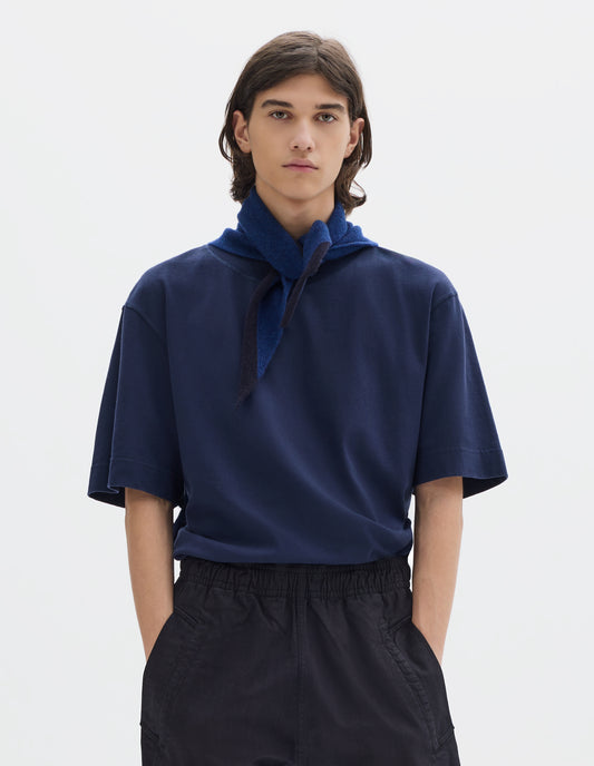 Cropped body image, front view of model wearing a blue wool triangle scarf with navy edge tied loosely on his neck. Styled with blue short sleeve jersey and dark navy jogger style trousers.