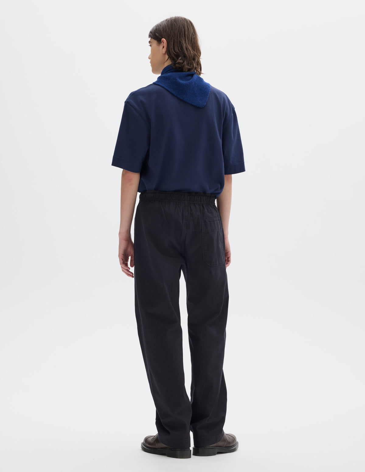Full body image, back view of model wearing relaxed dark blue joggers with elasticated waist and patch back pocket. Styled with a relaxed blue short sleeve t-shirt, blue and navy wool scout scarf and dark brown leather shoes.