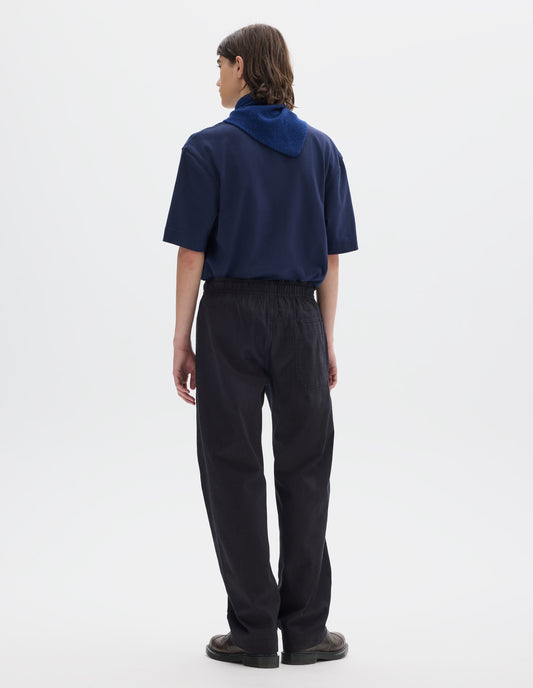 Full body image, back view of model wearing relaxed dark blue joggers with elasticated waist and patch back pocket. Styled with a relaxed blue short sleeve t-shirt, blue and navy wool scout scarf and dark brown leather shoes.