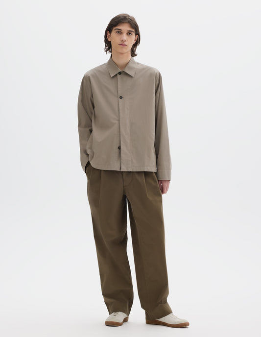 Full body image, front view of model wearing a relaxed boxy greyish beige overshirt with grown-on cuffs, stitch-through faced placket. Overshirt is worn half buttoned up, styled with khaki brown pleated trousers and light beige trainers.