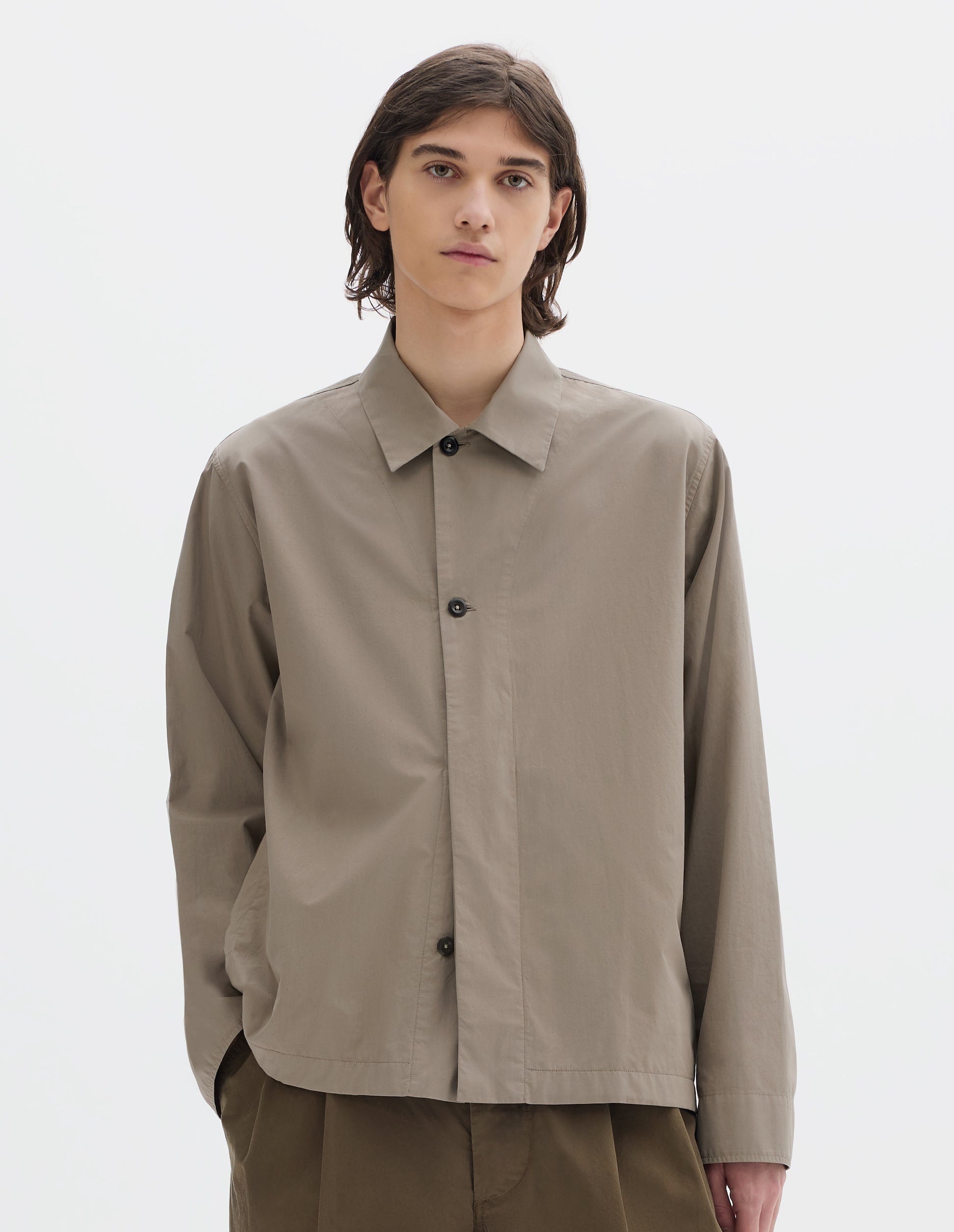 Cropped body image, front view of model wearing a relaxed boxy greyish beige overshirt with grown-on cuffs, and a stitch-through yoke. Overshirt is worn half buttoned up, styled with khaki brown pleated trousers.
