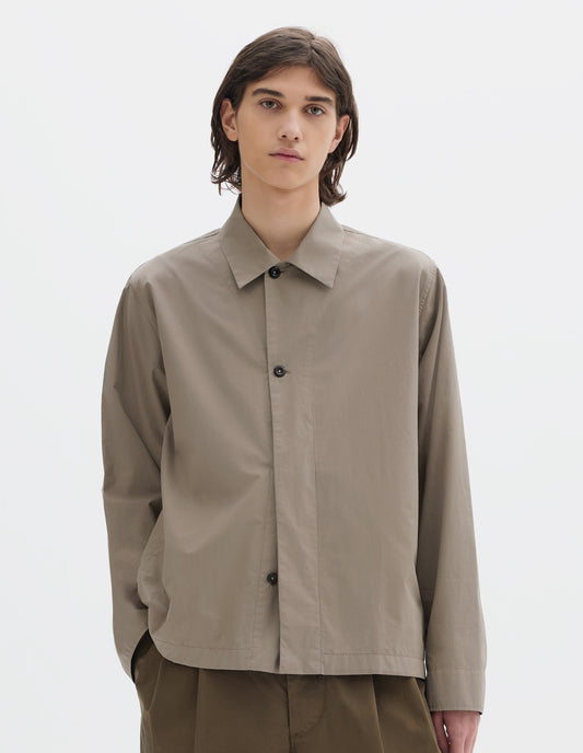 Cropped body image, front view of model wearing a relaxed boxy greyish beige overshirt with grown-on cuffs, and a stitch-through yoke. Overshirt is worn half buttoned up, styled with khaki brown pleated trousers.