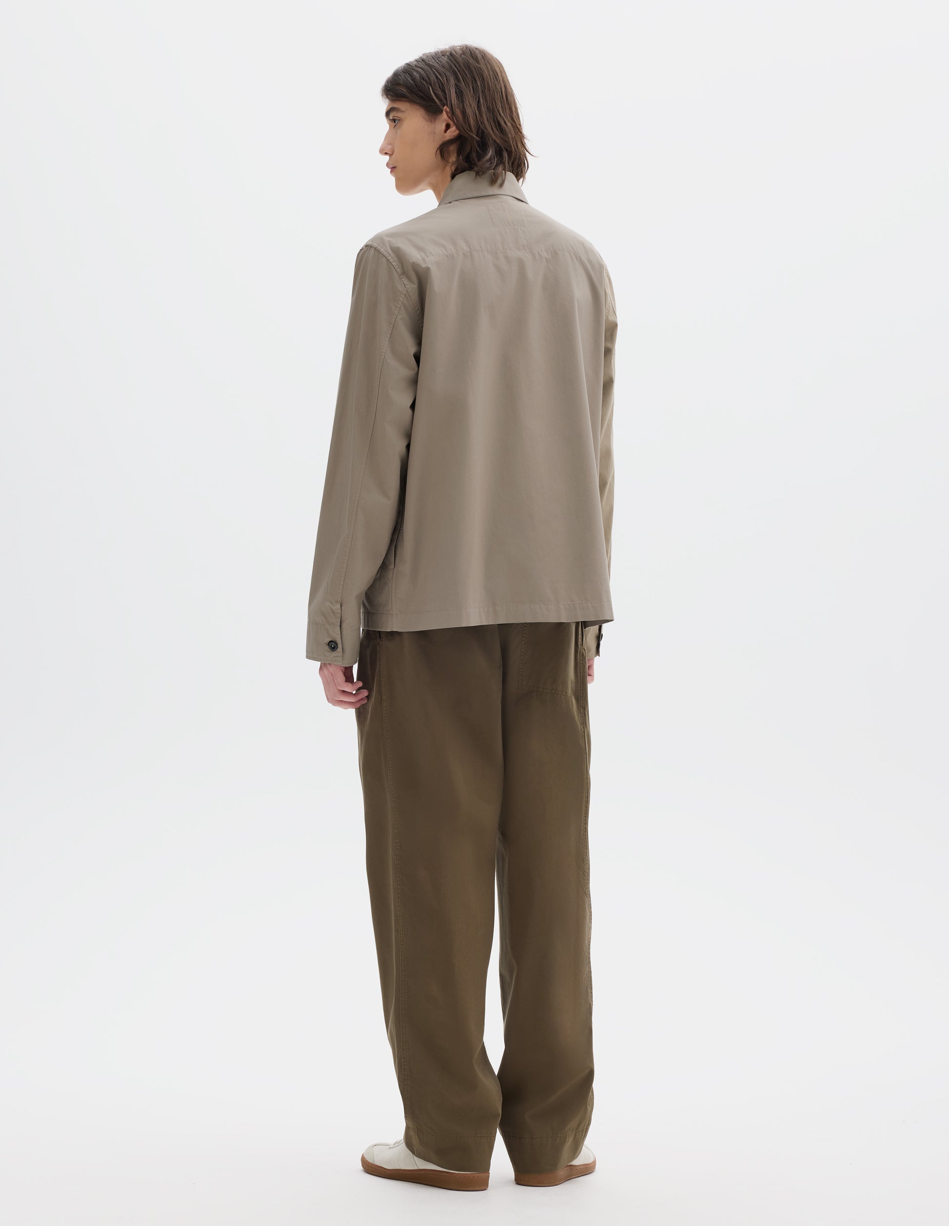 Full body image, back view of model wearing relaxed boxy greyish beige overshirt with grown-on cuffs and a stitch-through yoke. styled with khaki brown pleated trousers and light beige trainers.