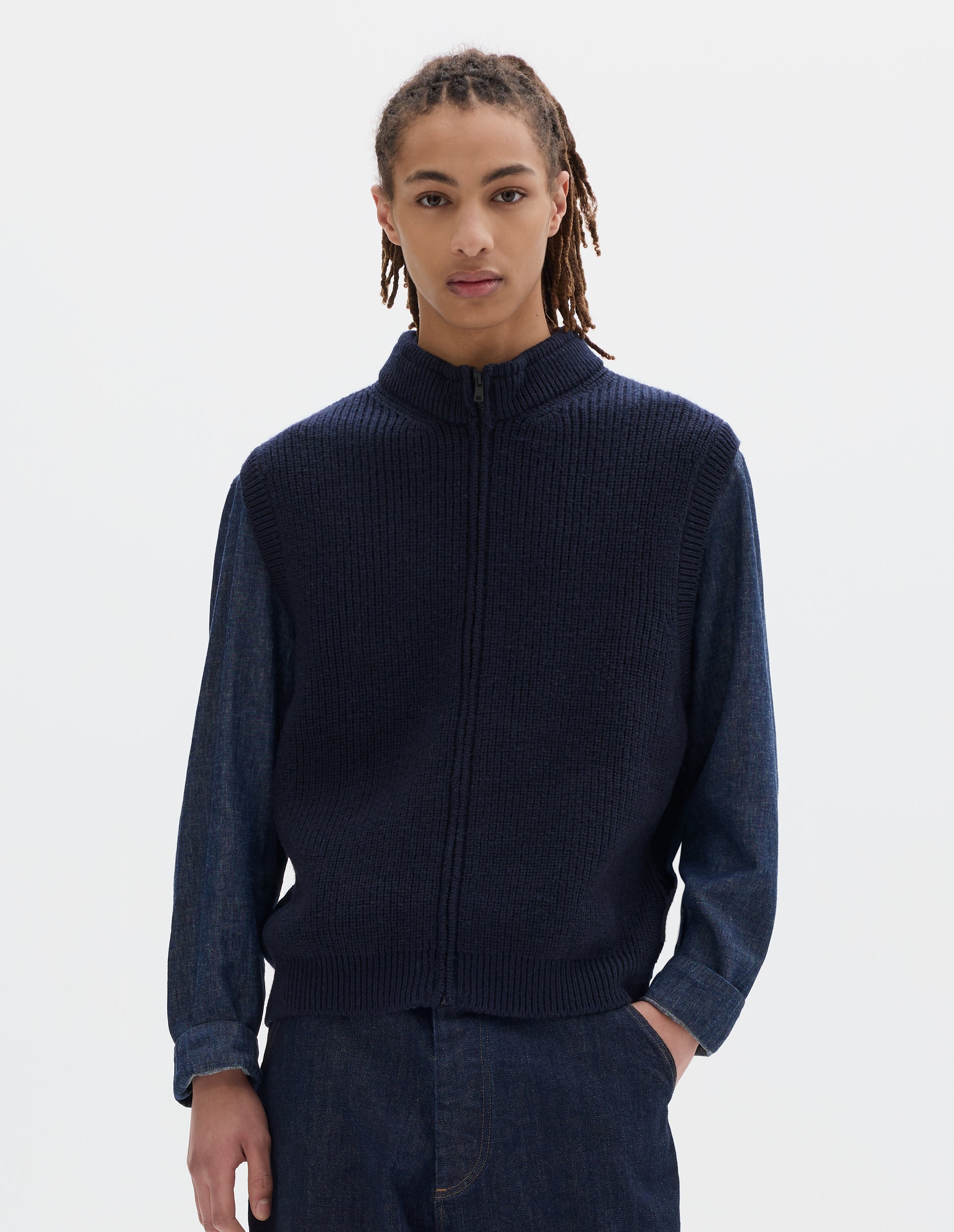 Cropped body image, front view of model wearing dark blue wool gilet, with rib neck, armholes and hem, and zip. Styled with dark grey shirt and blue jeans.