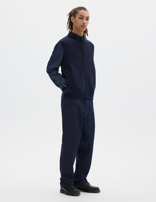 Full body image, front view of model wearing dark blue wool gilet, with rib neck, armholes and hem, and zip. Styled with dark grey shirt, blue jeans and black shoes.