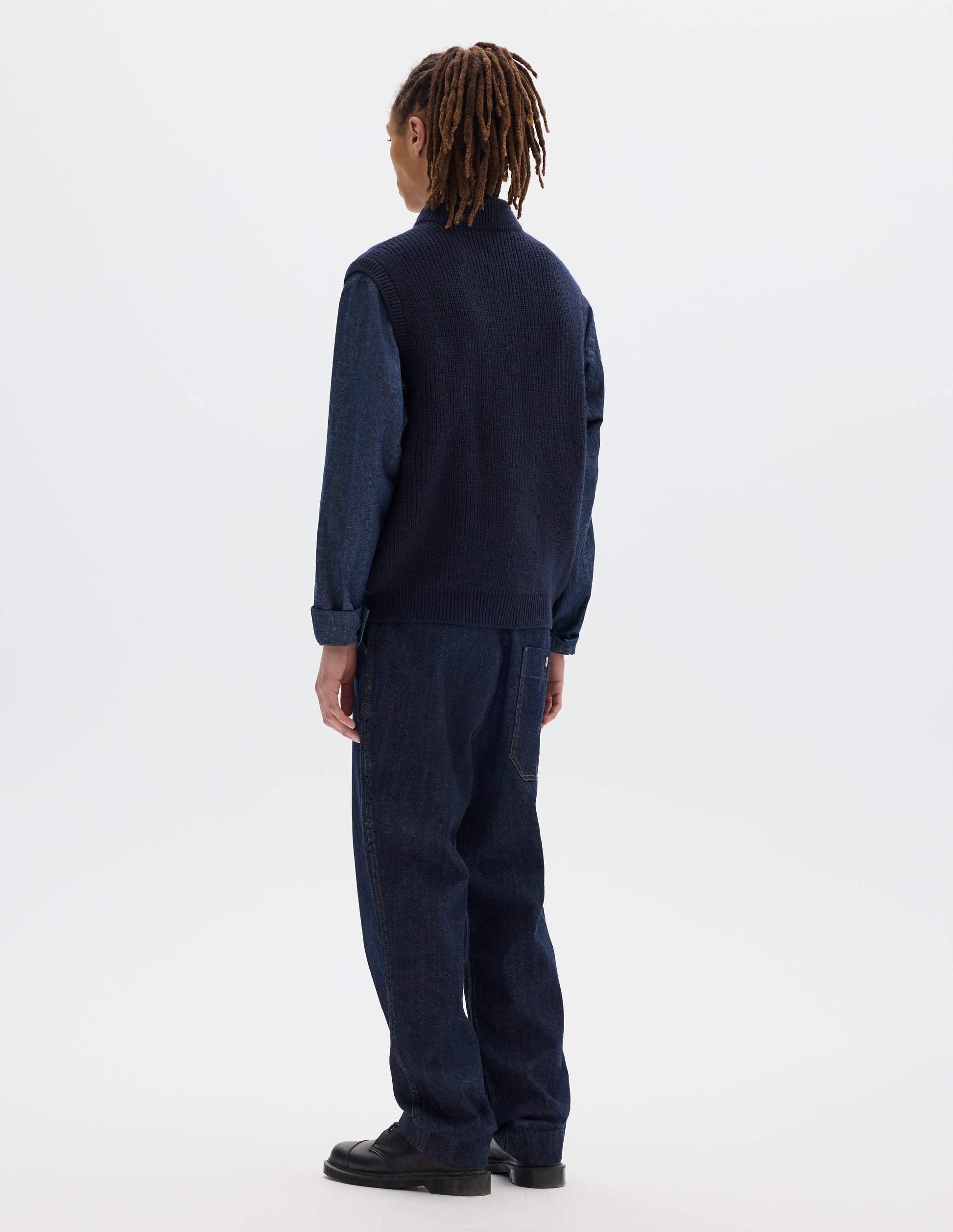 Full body image, back view of model wearing dark blue wool gilet, with rib neck, armholes and hem, and zip. Styled with dark grey shirt, blue jeans and black shoes.