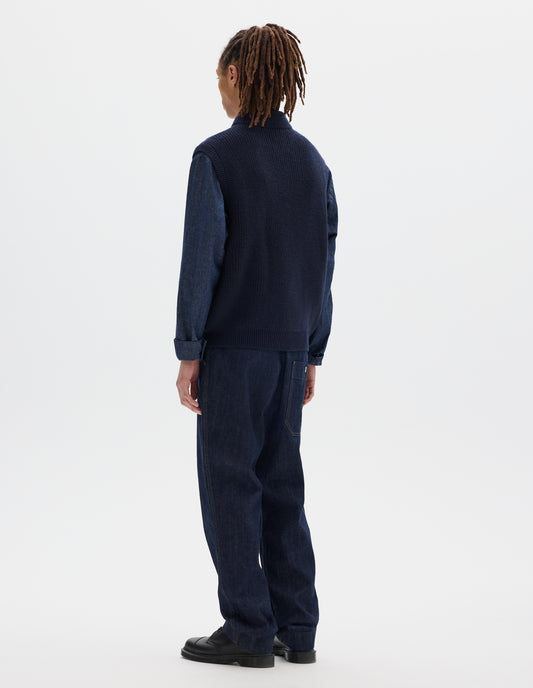 Full body image, back view of model wearing dark blue wool gilet, with rib neck, armholes and hem, and zip. Styled with dark grey shirt, blue jeans and black shoes.
