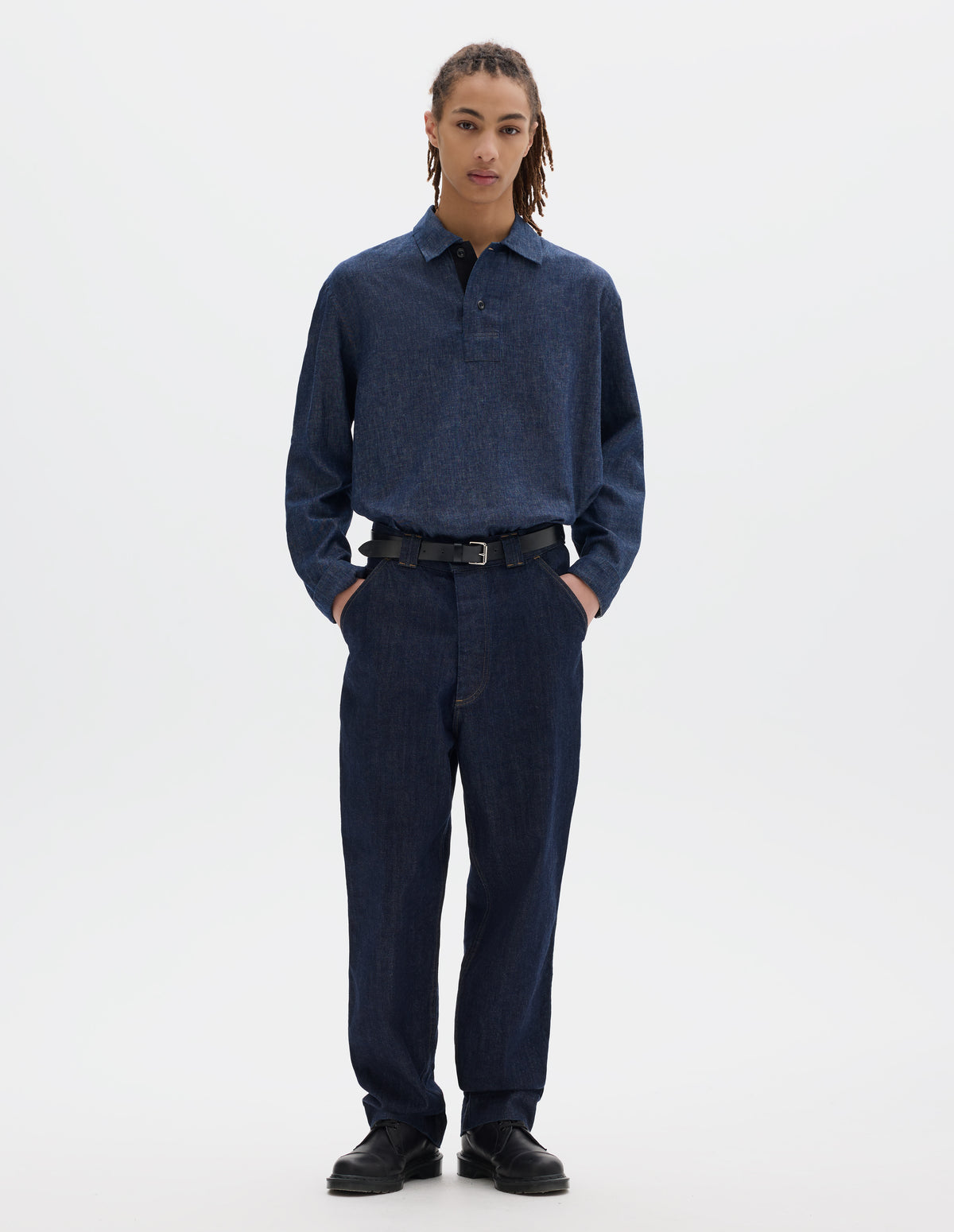 Full body image, front view of model wearing an indigo blue denim twill jeans with metal button fly, wide belt loops and slant pockets with ticket pocket detailing. Styled with a blue cotton linen long sleeve polo shirt, black leather belt and shoes.
