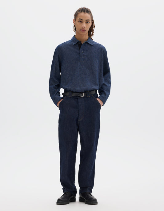 Full body image, front view of model wearing an indigo blue denim twill jeans with metal button fly, wide belt loops and slant pockets with ticket pocket detailing. Styled with a blue cotton linen long sleeve polo shirt, black leather belt and shoes.