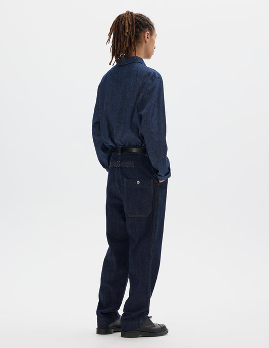 Full body image, back view of model wearing an indigo blue denim twill jeans with metal cinch on back, wide belt loops and dropped metal button through patch back pocket. Styled with a blue cotton linen long sleeve polo shirt, black leather belt and shoes.