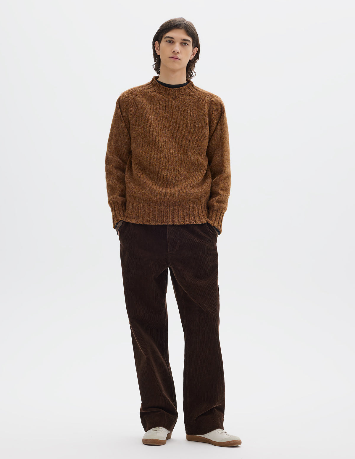 Full body image, front view of model wearing a brown cotton corduroy trousers with slanted side pockets. Styled with orangey brown long sleeve wool jumper, with contrast ribbing on neckline, cuffs and hem and off white suede trainers with gum sole.