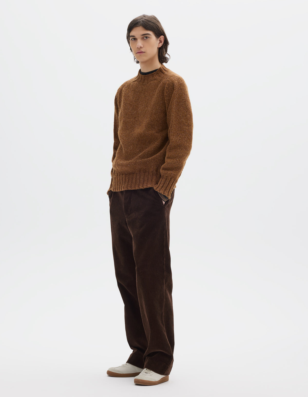 Full body image, front view of model wearing orangey brown long sleeve wool jumper, with contrast ribbing on neckline, cuffs and hem. Styled with brown cotton corduroy trousers and white trainers with gum sole.
