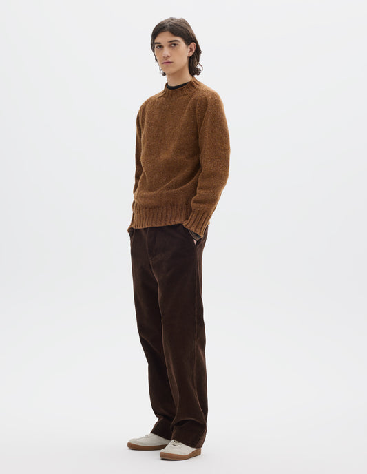 Full body image, front view of model wearing orangey brown long sleeve wool jumper, with contrast ribbing on neckline, cuffs and hem. Styled with brown cotton corduroy trousers and white trainers with gum sole.