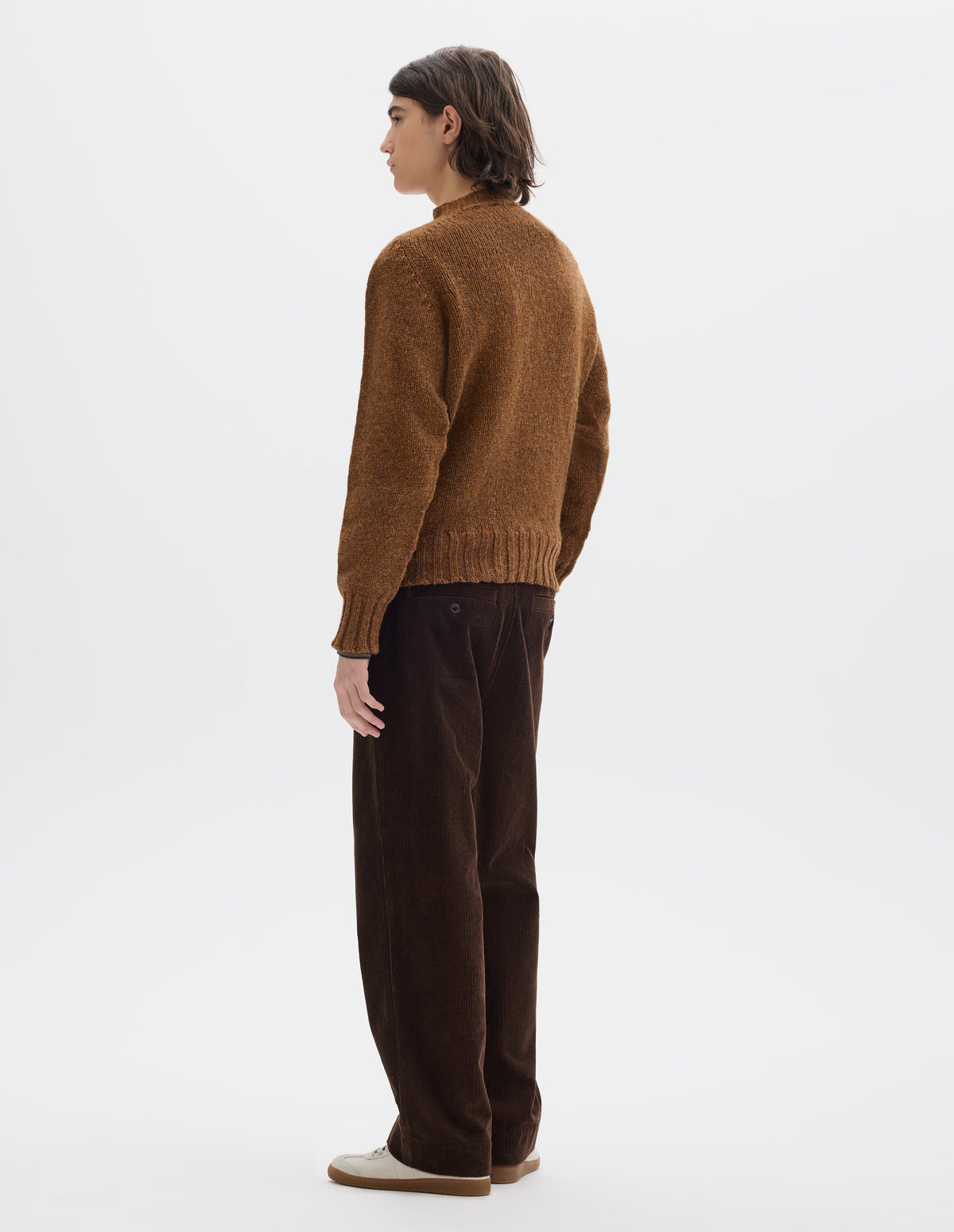 Full body image, back view of model wearing orangey brown long sleeve wool jumper, with contrast ribbing on neckline, cuffs and hem. Styled with brown cotton corduroy trousers and white trainers with gum sole.