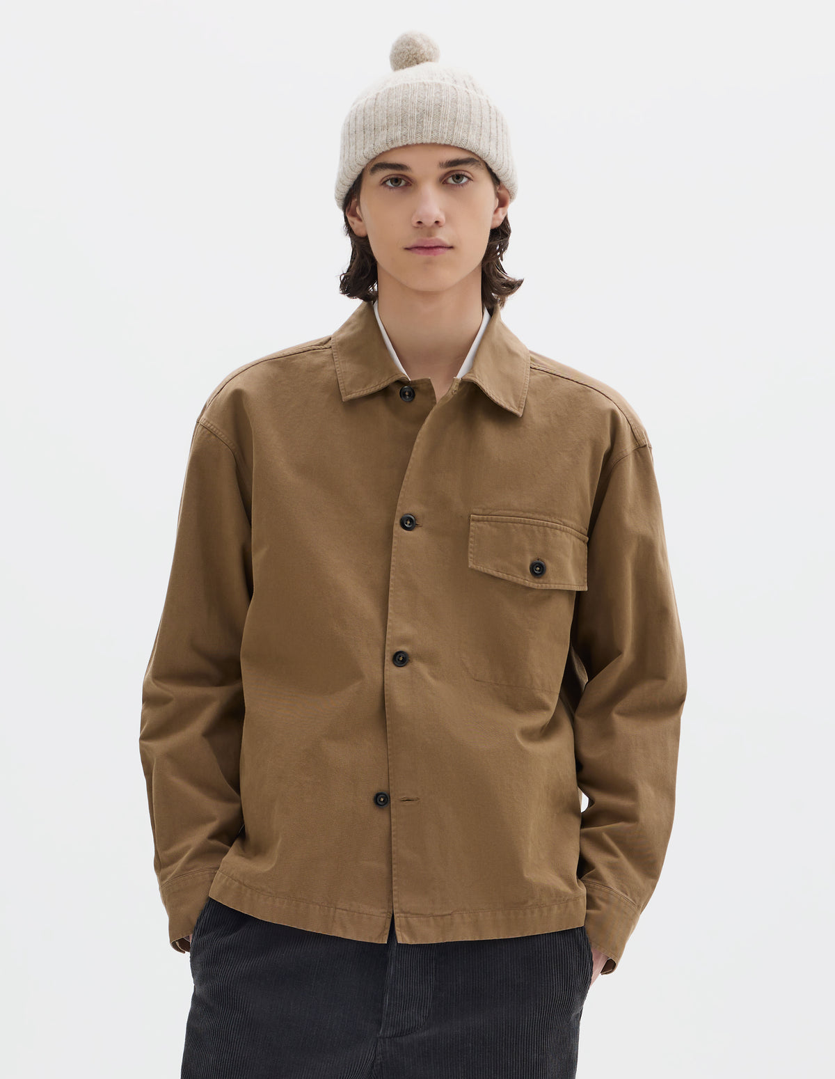 Cropped body image, front view of model wearing a greyish beige knitted wool bobble hat with a rib turn up. Styled with a light brown cotton overshirt and dark grey cord trousers.