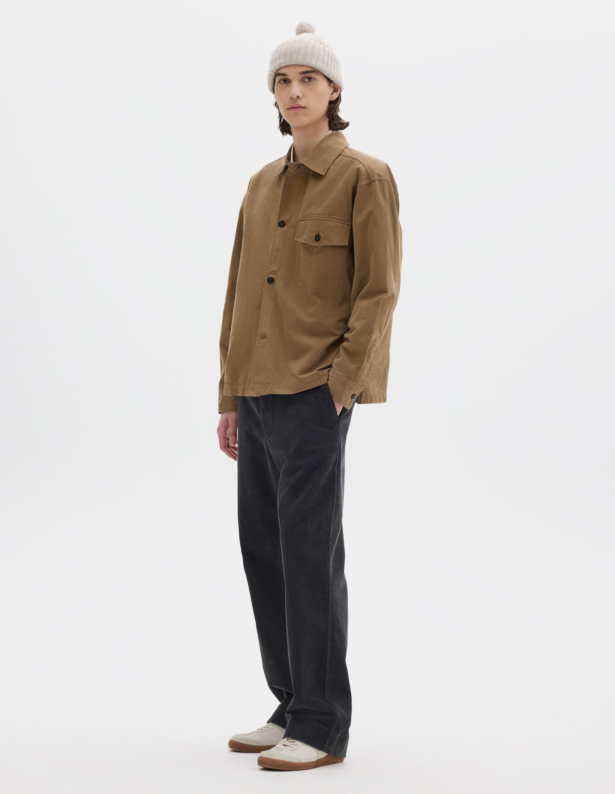 Full body image, front view of model wearing brownish green cotton overshirt, with a grown-on, button-fastening placket, spread collar, and welt chest pockets with flaps and button closure, finished with shirt cuffs. Styled with off white bobble beanie and dark grey corduroy trousers, with white trainers with gum sole.