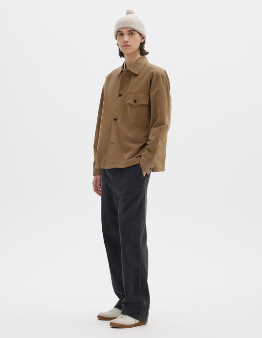 Full body image, front view of model wearing brownish green cotton overshirt, with a grown-on, button-fastening placket, spread collar, and welt chest pockets with flaps and button closure, finished with shirt cuffs. Styled with off white bobble beanie and dark grey corduroy trousers, with white trainers with gum sole.