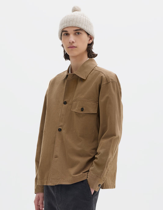 Cropped body image, front view of model wearing brownish green cotton overshirt, with a grown-on, button-fastening placket, spread collar, and welt chest pockets with flaps and button closure, finished with shirt cuffs. Styled with off white bobble beanie and dark grey corduroy trousers.