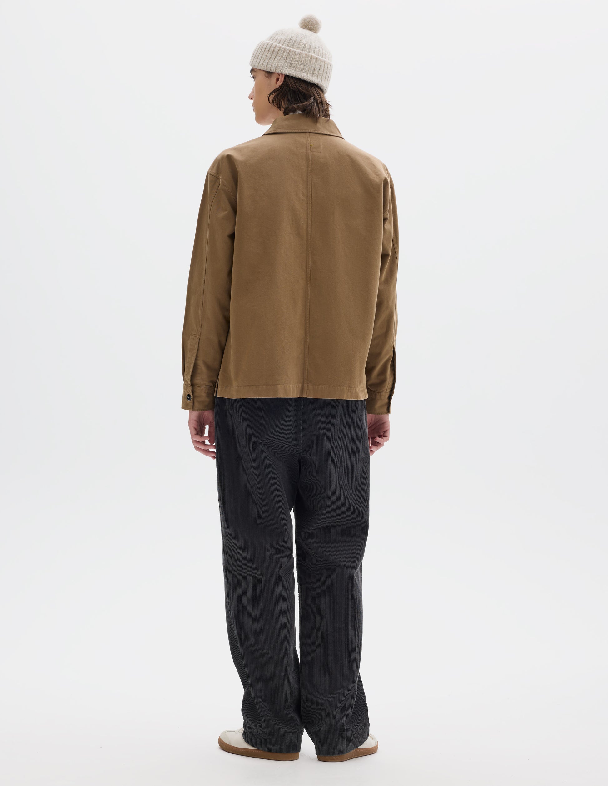 Full body image, back view of model wearing brownish green cotton overshirt, with button closure shirt cuffs and centre back seam. Styled with off white bobble beanie and dark grey corduroy trousers, with white trainers with gum sole.