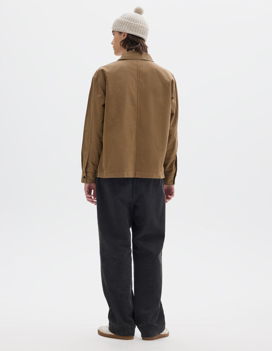 Full body image, back view of model wearing brownish green cotton overshirt, with button closure shirt cuffs and centre back seam. Styled with off white bobble beanie and dark grey corduroy trousers, with white trainers with gum sole.
