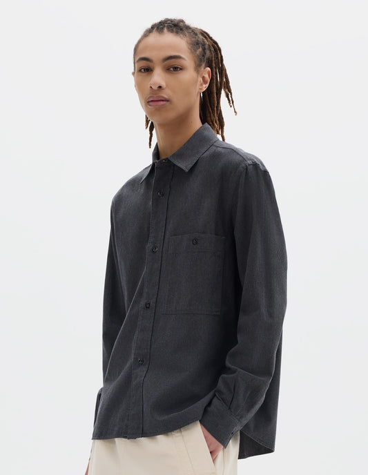 Cropped body image, front view of model wearing dark grey cotton chambray shirt with a dropped shoulder, raised placket and button through chest patch pocket. Styled with off white joggers.