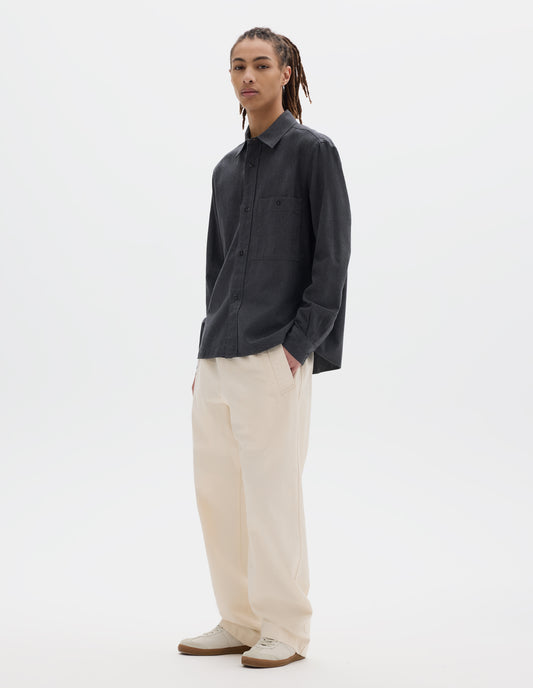 Full body image, front view of model wearing dark grey cotton chambray shirt with a dropped shoulder, raised placket and button through chest patch pocket. Styled with off white joggers and white trainers with gum sole. 