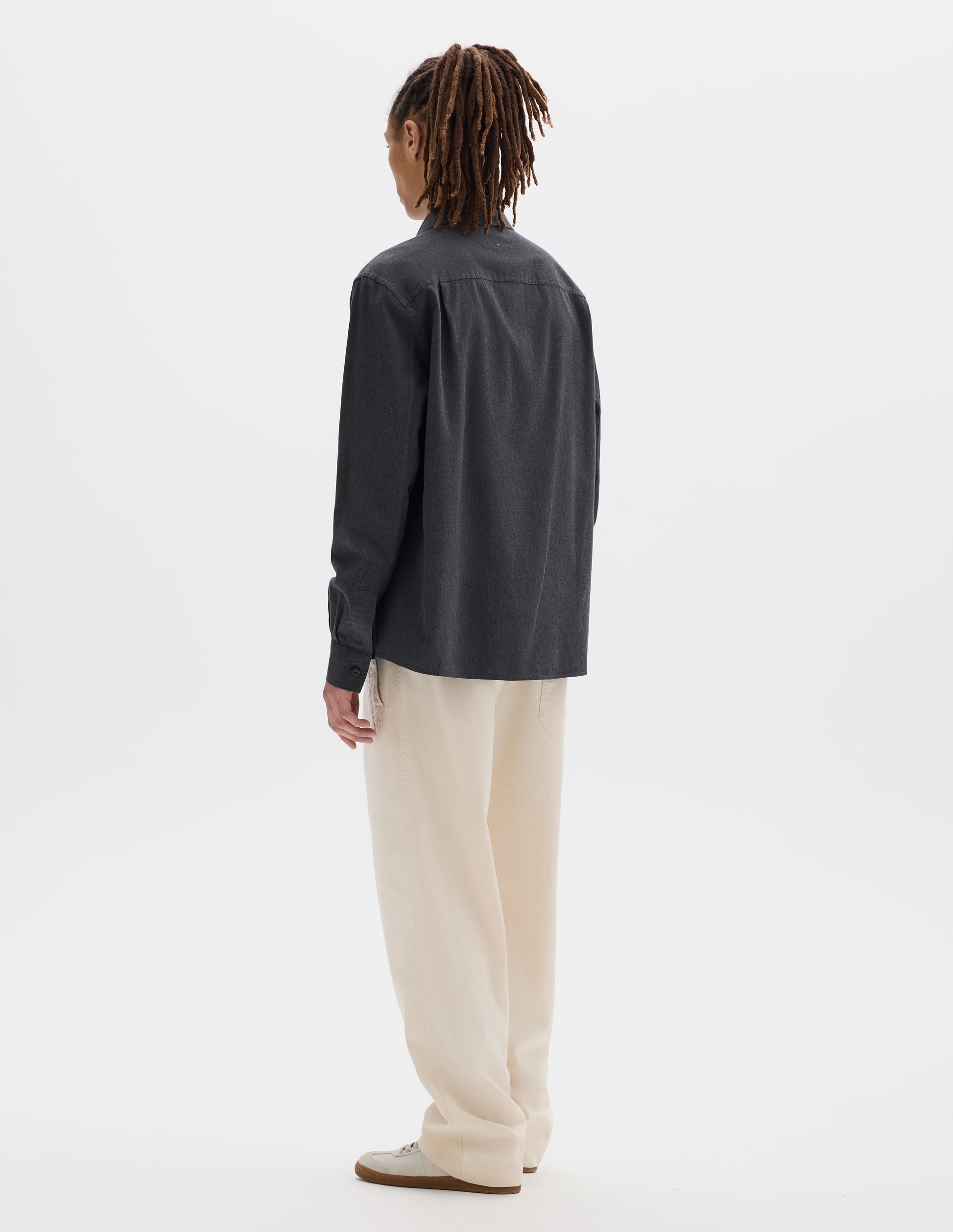 Full body image, back view of model wearing dark grey cotton chambray shirt with a dropped shoulder and pleats on back yoke. Styled with off white joggers and white trainers with gum sole. 