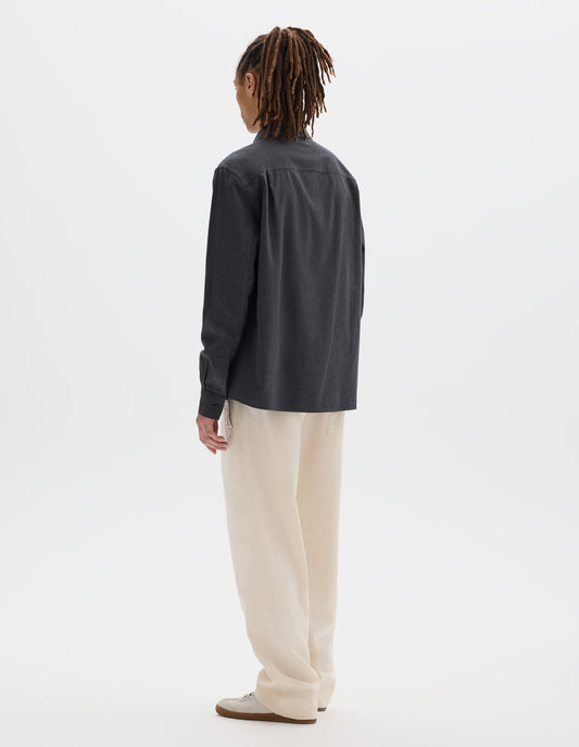 Full body image, back view of model wearing dark grey cotton chambray shirt with a dropped shoulder and pleats on back yoke. Styled with off white joggers and white trainers with gum sole. 