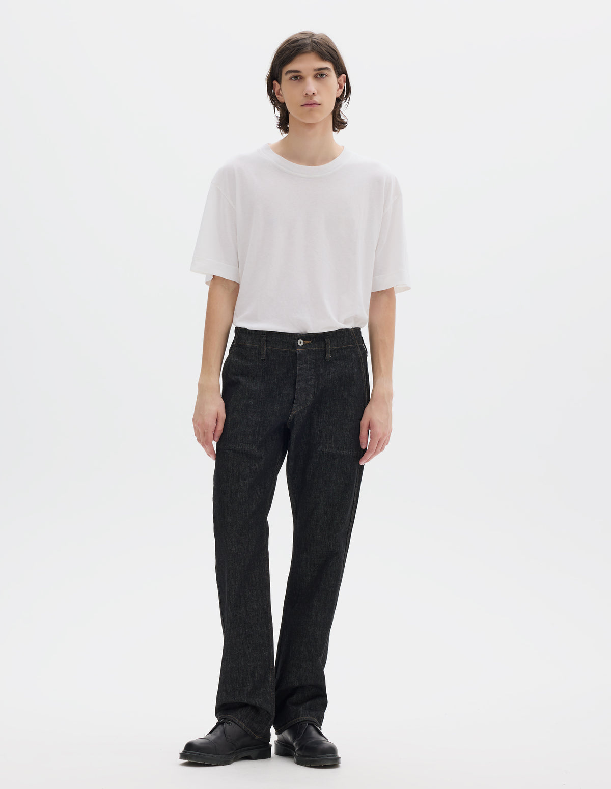 Full body image, front view of model wearing black denim twill jeans, with metal button fly, grown on waistband with belt loops and stitch-through slant pockets. Styled with white short sleeve T shirt and black leather shoes.