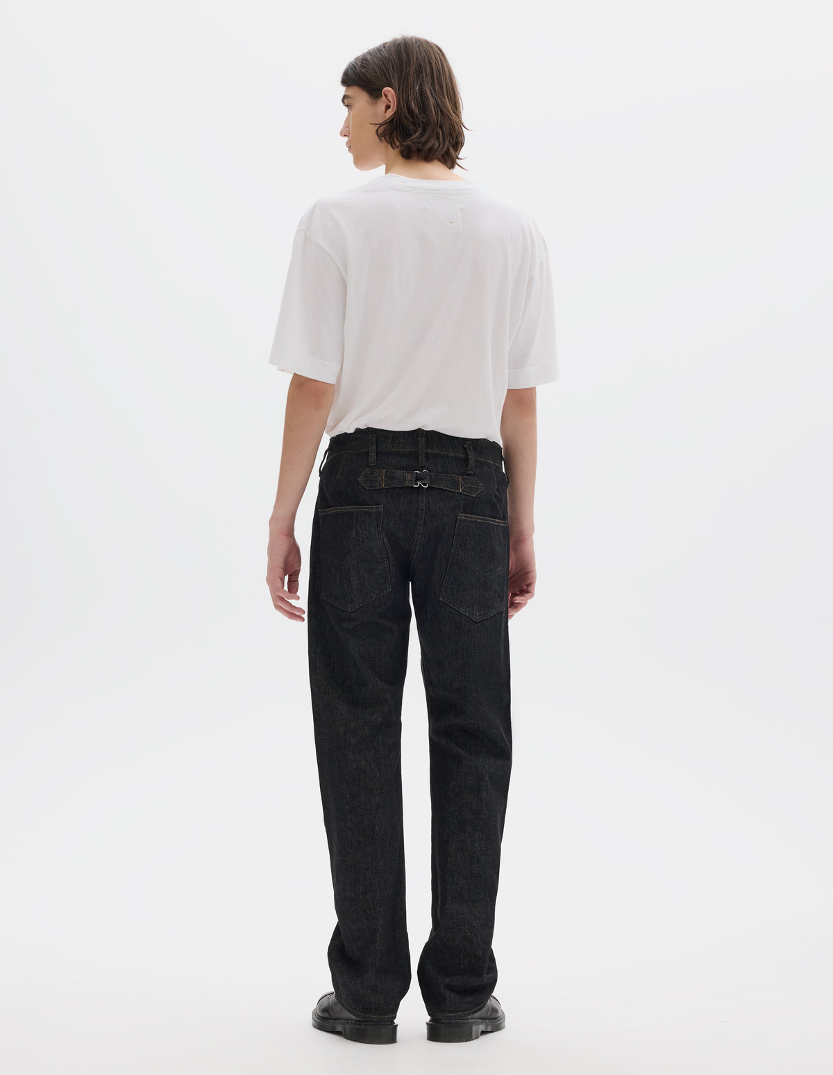 Full body image, back view of model wearing black denim twill jeans, with stitch-through waistband with belt loops, cinch back detail with metal buckle and patch back pockets. Styled with white short sleeve T shirt and black leather shoes.