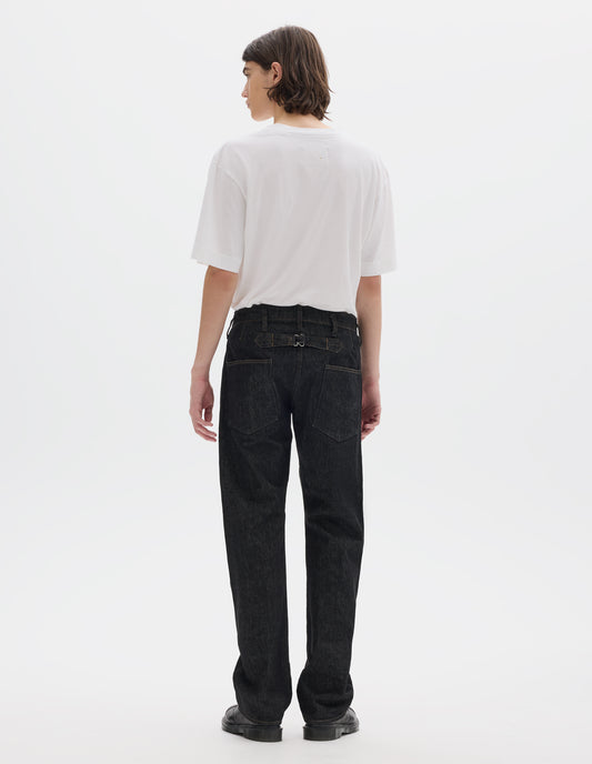 Full body image, back view of model wearing black denim twill jeans, with stitch-through waistband with belt loops, cinch back detail with metal buckle and patch back pockets. Styled with white short sleeve T shirt and black leather shoes.