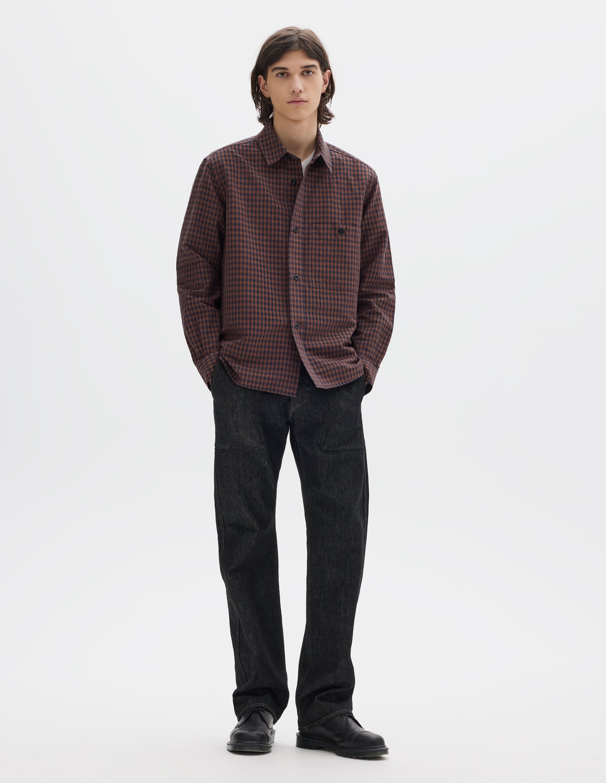 Full body image, front view of model wearing brown and dark blue check cotton linen shirt, with button-through chest patch pocket and dark buttons. Styled with white t shirt, black jeans and black shoes.