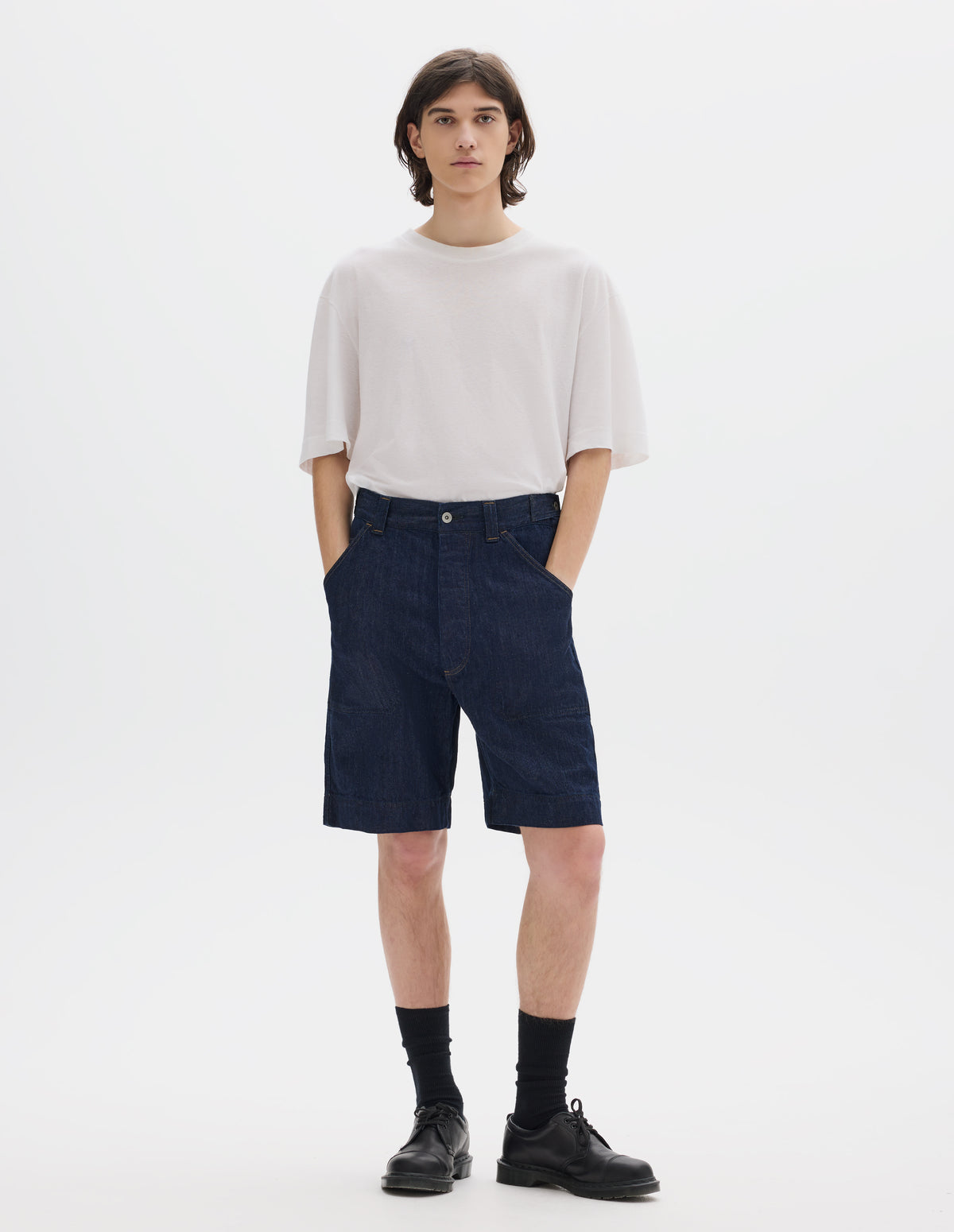 Full body image, front view of model wearing dark blue relaxed denim twill shorts with metal button tab waist adjuster, metal button fly, belt loops and slant stitch-through pockets. Styled with white short sleeve linen cotton T shirt, black socks and blacl leather shoes.