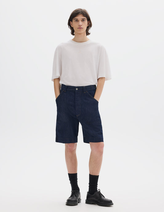 Full body image, front view of model wearing dark blue relaxed denim twill shorts with metal button tab waist adjuster, metal button fly, belt loops and slant stitch-through pockets. Styled with white short sleeve linen cotton T shirt, black socks and blacl leather shoes.
