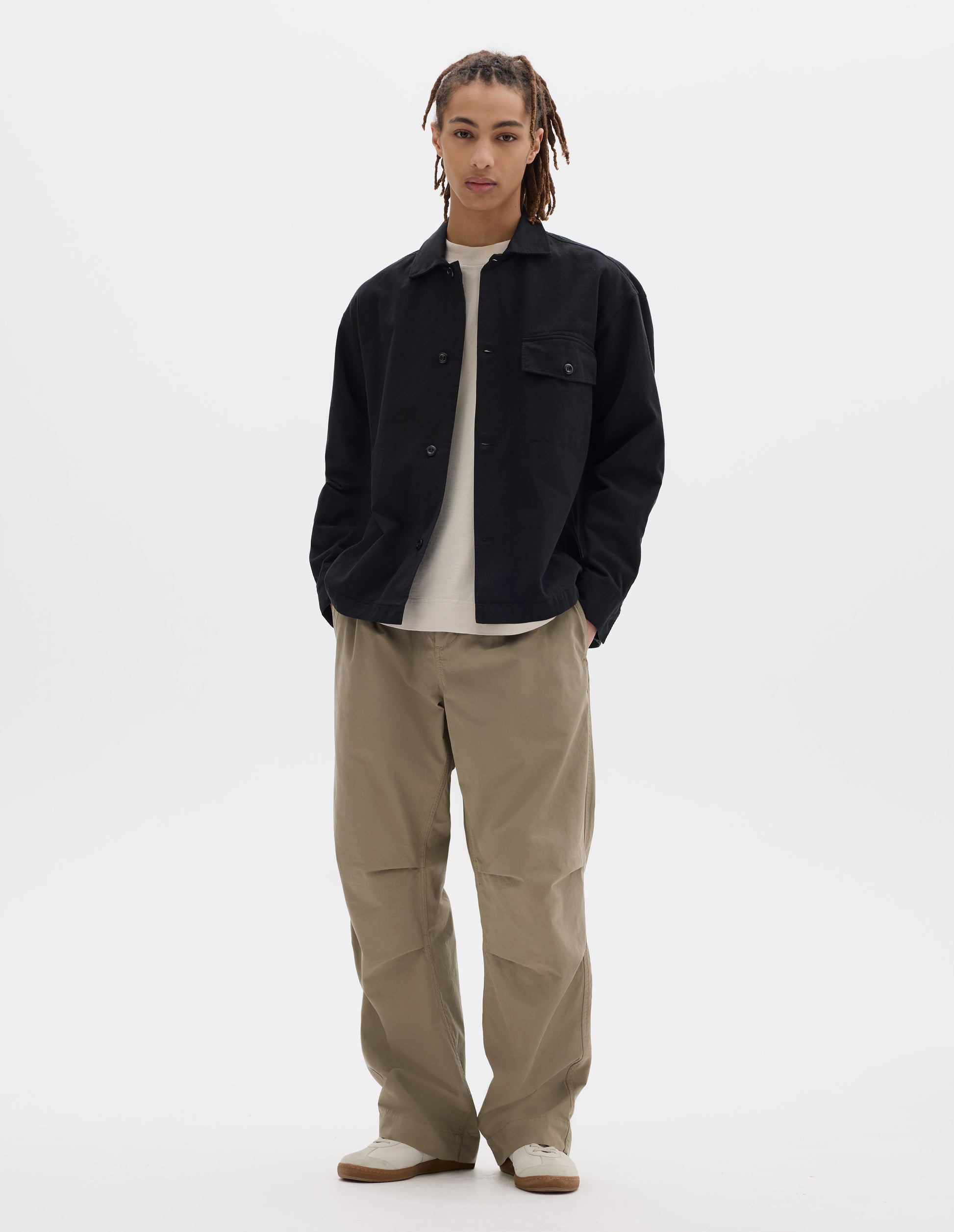 Full body image, front view of model wearing black cotton overshirt, with a grown-on, button-fastening placket, spread collar, and welt chest pockets with flaps and button closure, finished with shirt cuffs. Styled over off white t-shirt, beige trousers and white trainers with gum sole.