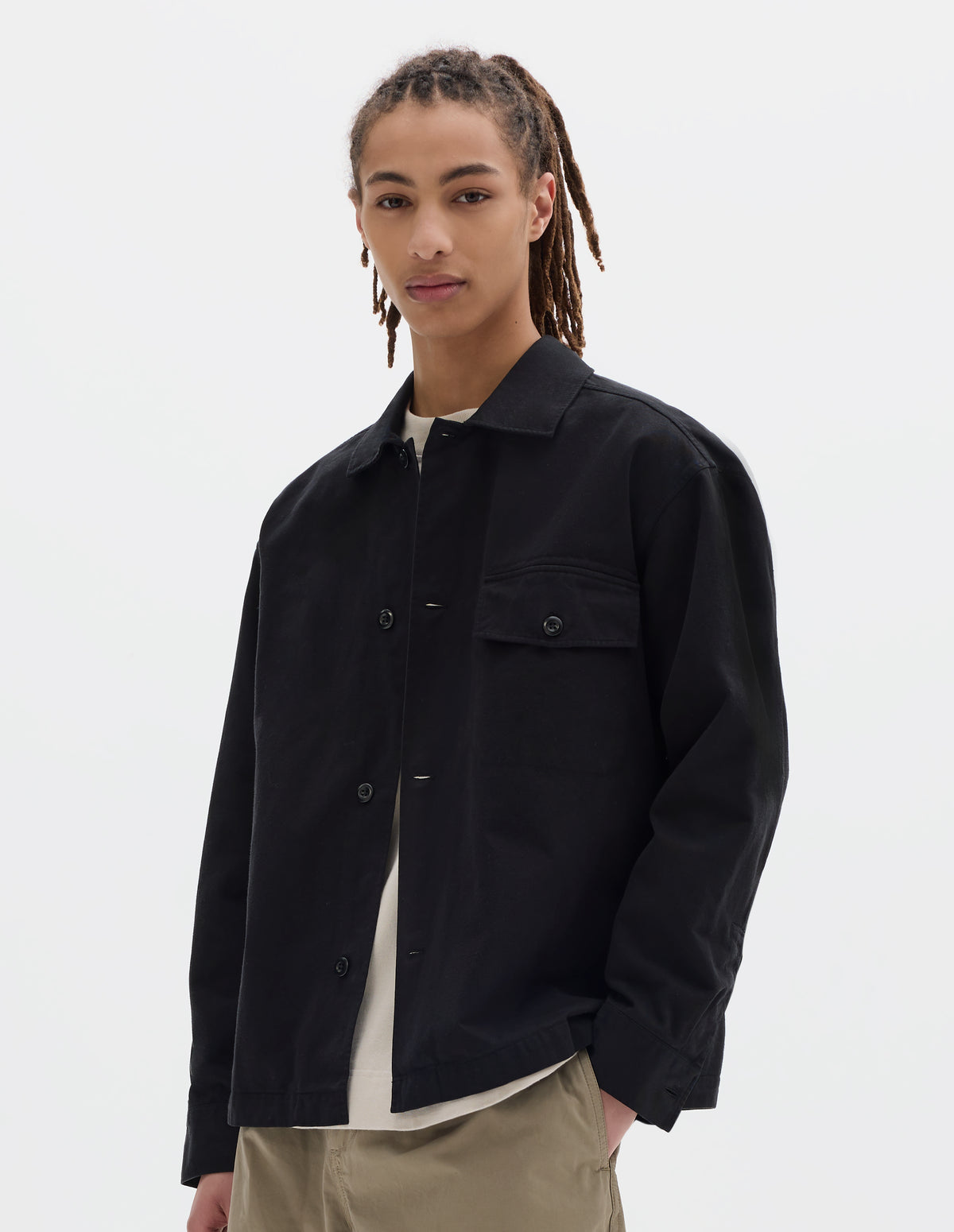 Cropped body image, front view of model wearing black cotton overshirt, with a grown-on, button-fastening placket, spread collar, and welt chest pockets with flaps and button closure, finished with shirt cuffs. Styled over off white t-shirt with beige trousers.